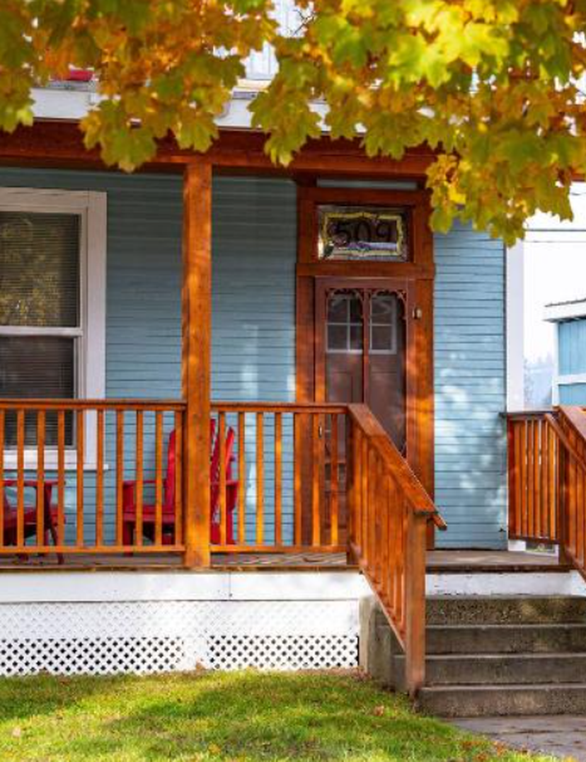 A cozy blue house with a wooden porch, red chairs, and autumn leaves on a tree, creating a warm and inviting atmosphere.