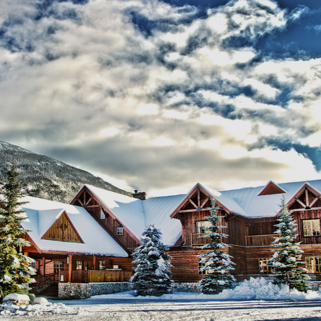 Rustic wooden lodge surrounded by snow-covered trees and mountains, under a vibrant blue sky with scattered clouds.