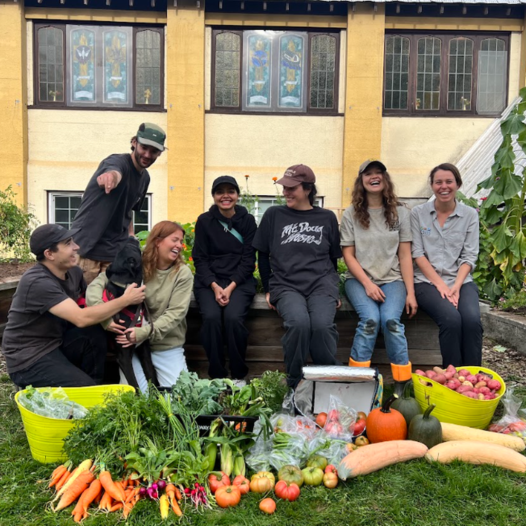 A group of people sitting outdoors, smiling, with baskets of fresh vegetables and fruits in front of them, near a garden and building.