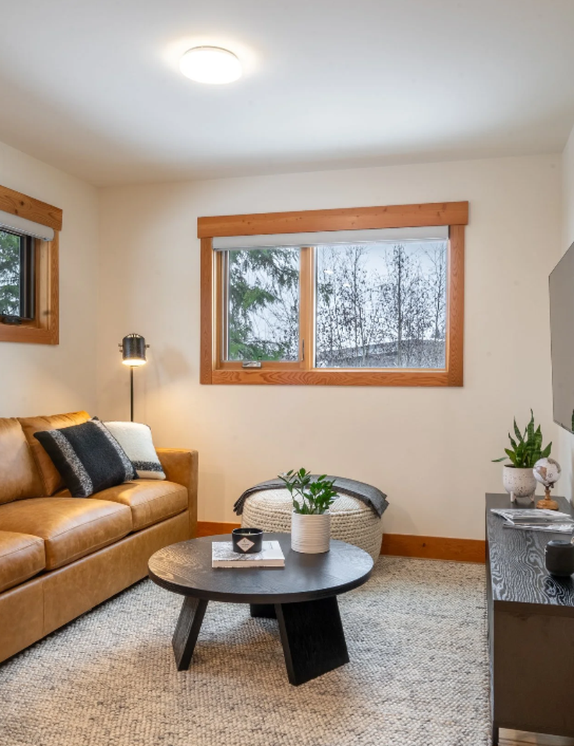 Cozy living room with tan leather sofa, round black coffee table, wall-mounted TV, wood-trim windows, pouf and potted plants.