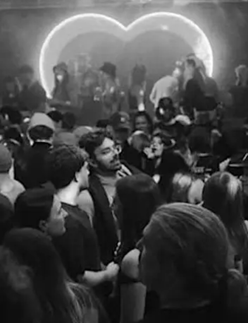 A crowded club party in black and white features people dancing under dim lighting, with a large illuminated heart in the background.