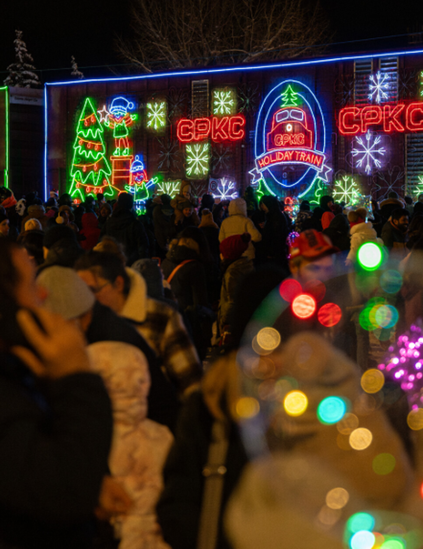 Crowd gathered at night in front of a brightly lit train with colorful holiday-themed neon decorations and snowflake designs.