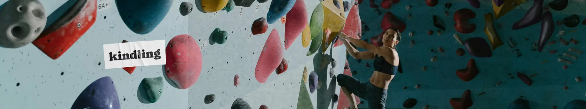 A person climbs a colorful indoor bouldering wall, surrounded by various shaped holds. The word "kindling" dispensary logo