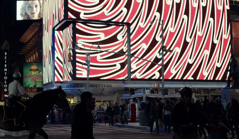 Photo of a busy downtown street at night. Electric billboards are filled with geometric designs in black, white and red.