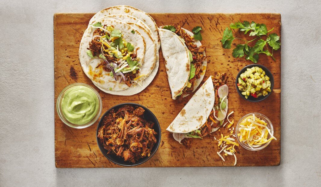 Tacos avec porc effiloché, sauce à l'avocat, fromage et salsa de maïs sur une planche en bois, garnis de coriandre.