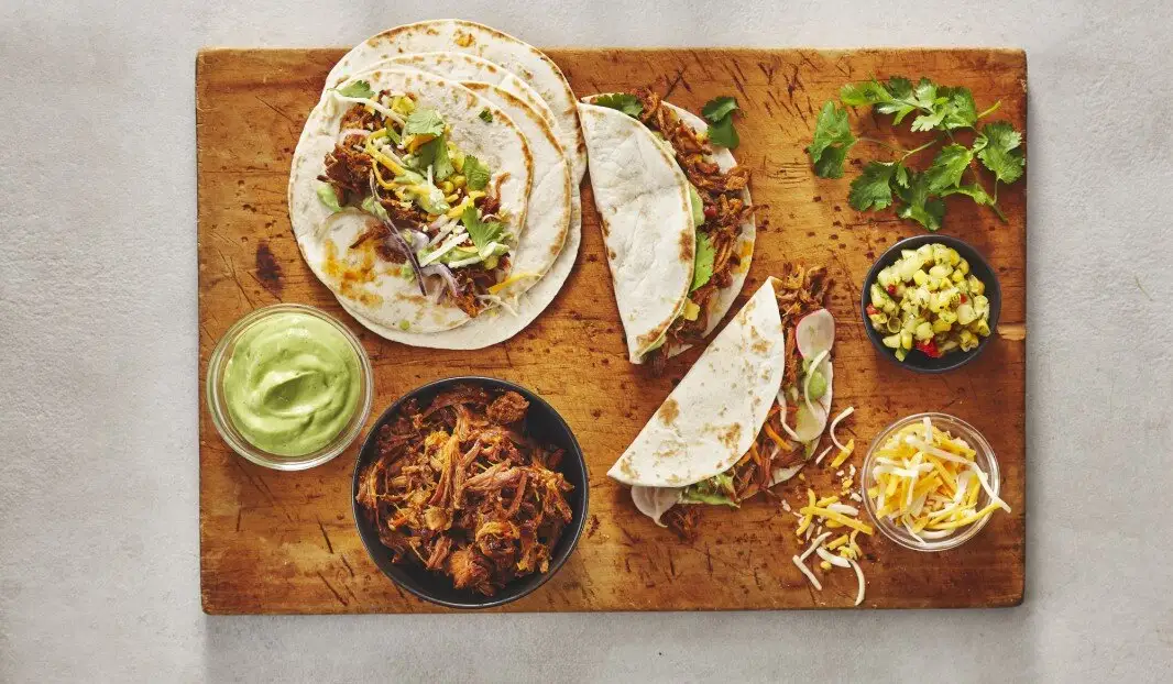 Tacos avec porc effiloché, sauce à l'avocat, fromage et salsa de maïs sur une planche en bois, garnis de coriandre.