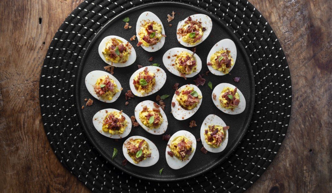 Quinze œufs mimosa garnis de bacon et herbes sur une assiette noire.