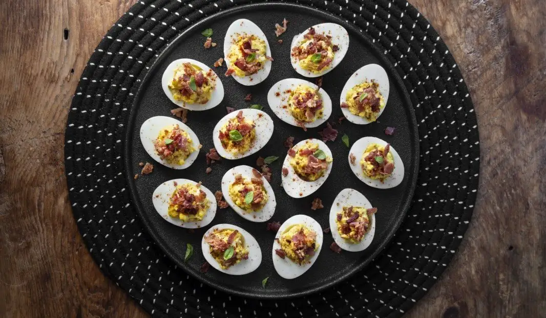 Quinze œufs mimosa garnis de bacon et herbes sur une assiette noire.