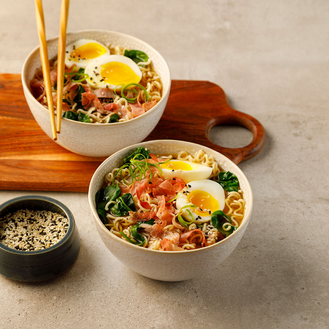 Deux bols blancs de ramens au jambon et oeuf mollet et un bol de graines de sésame noires et blanches sur un comptoir gris