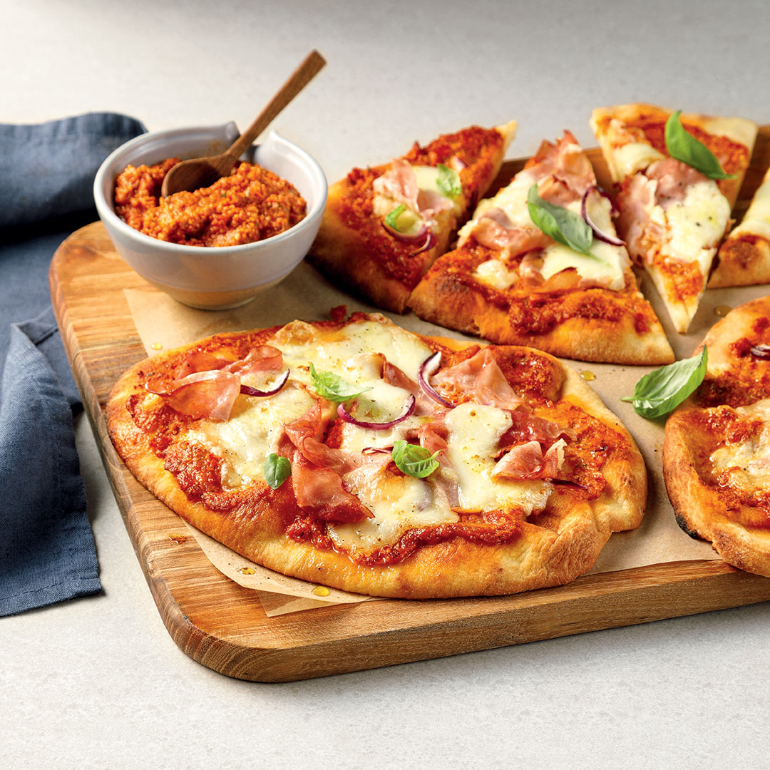 Pains plats au jambon, burrata et pesto servis sur une planche en bois et un bol de pesto