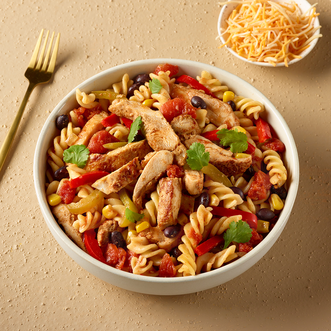 Pâtes à la mexicaine avec lanières de haut de cuisse de poulet, poivrons, maïs, haricots noirs et coriandre servies dans un bol blanc avec une fourchette or et un bol de fromage râpé