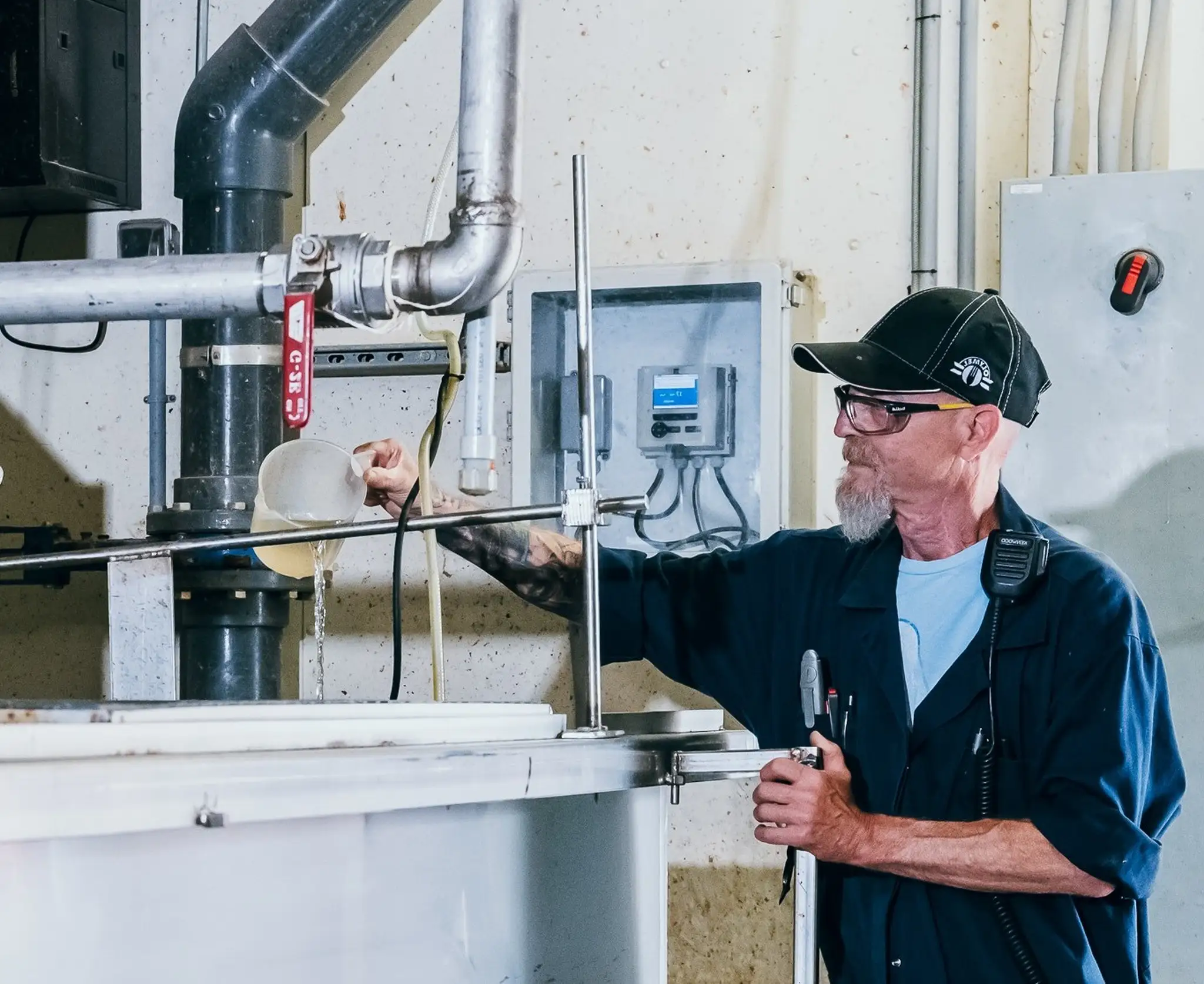 Homme portant une casquette noire et des lunettes, travaillant avec de l’équipement industriel, tenant un outil et versant un liquide dans un grand réservoir métallique.