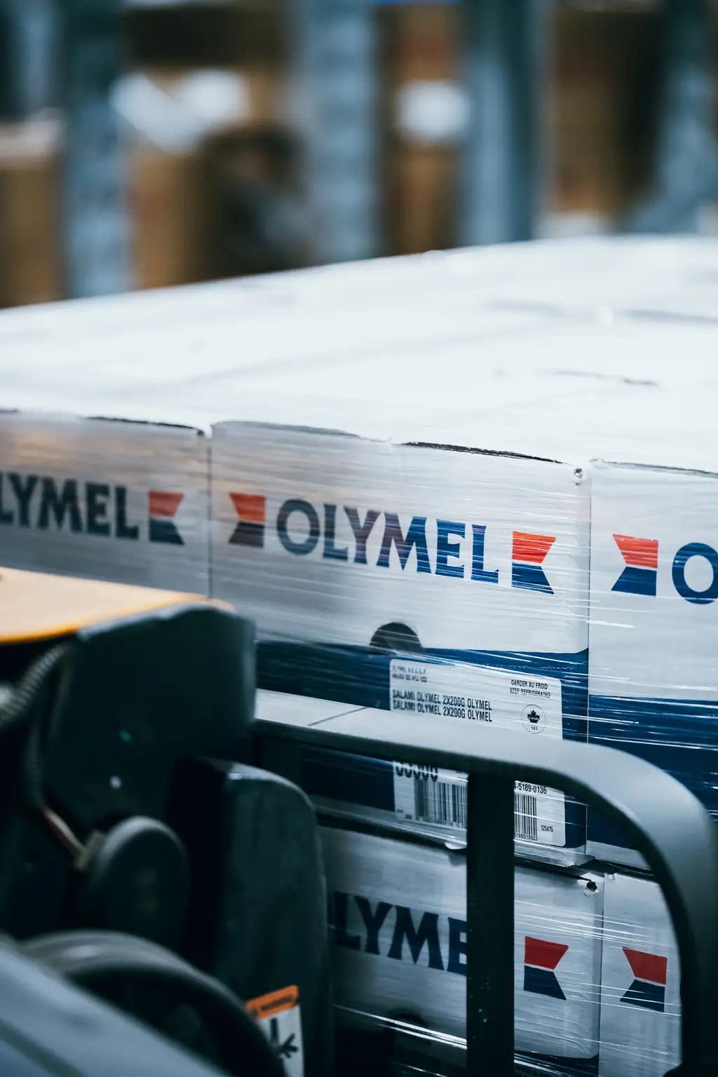 Des boîtes étiquetées "OLYMEL" empilées sur une palette, avec un chariot élévateur partiellement visible dans un entrepôt.