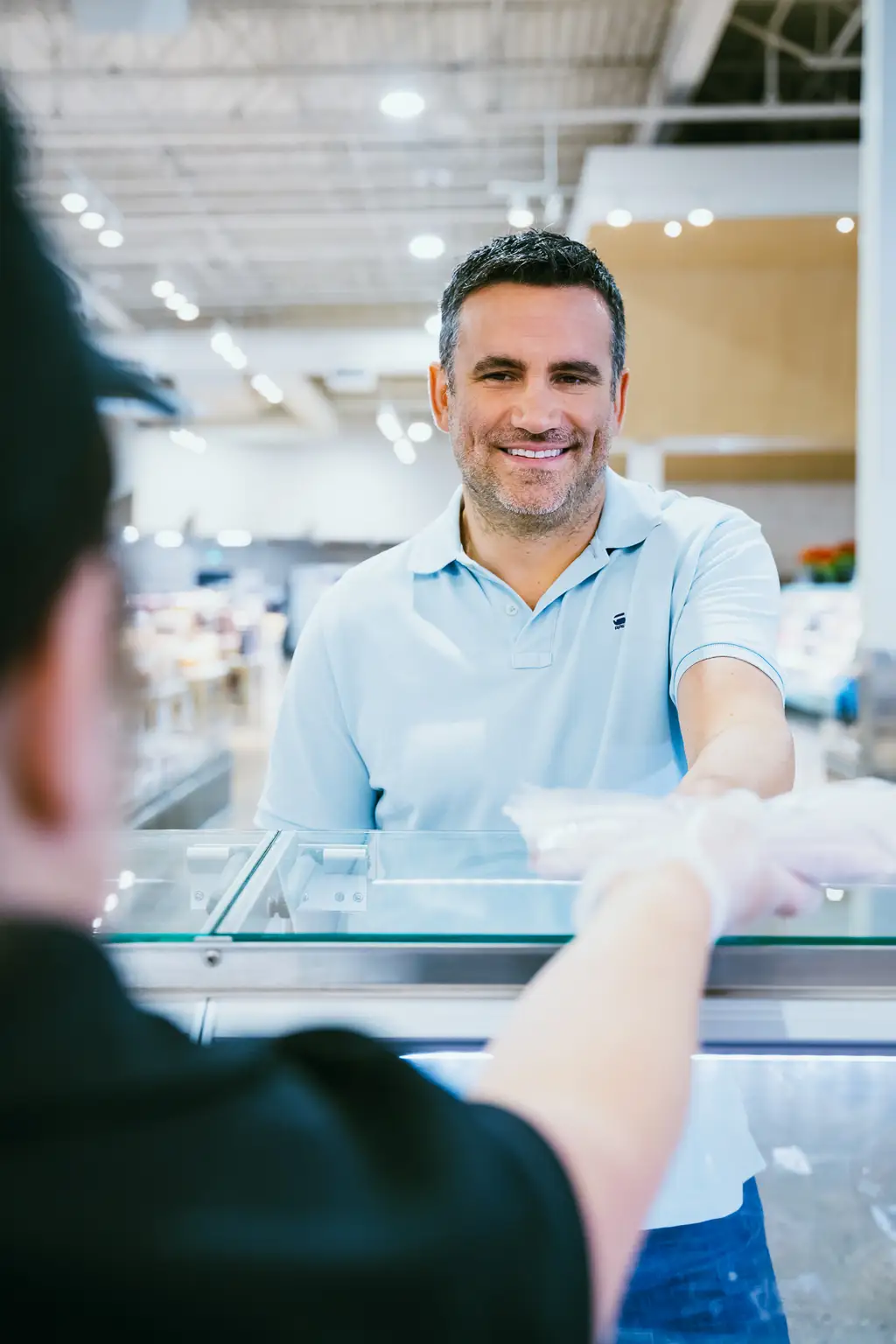 Un homme portant une chemise bleu pâle sourit en recevant un article emballé d’un employé ganté au comptoir d’un magasin.