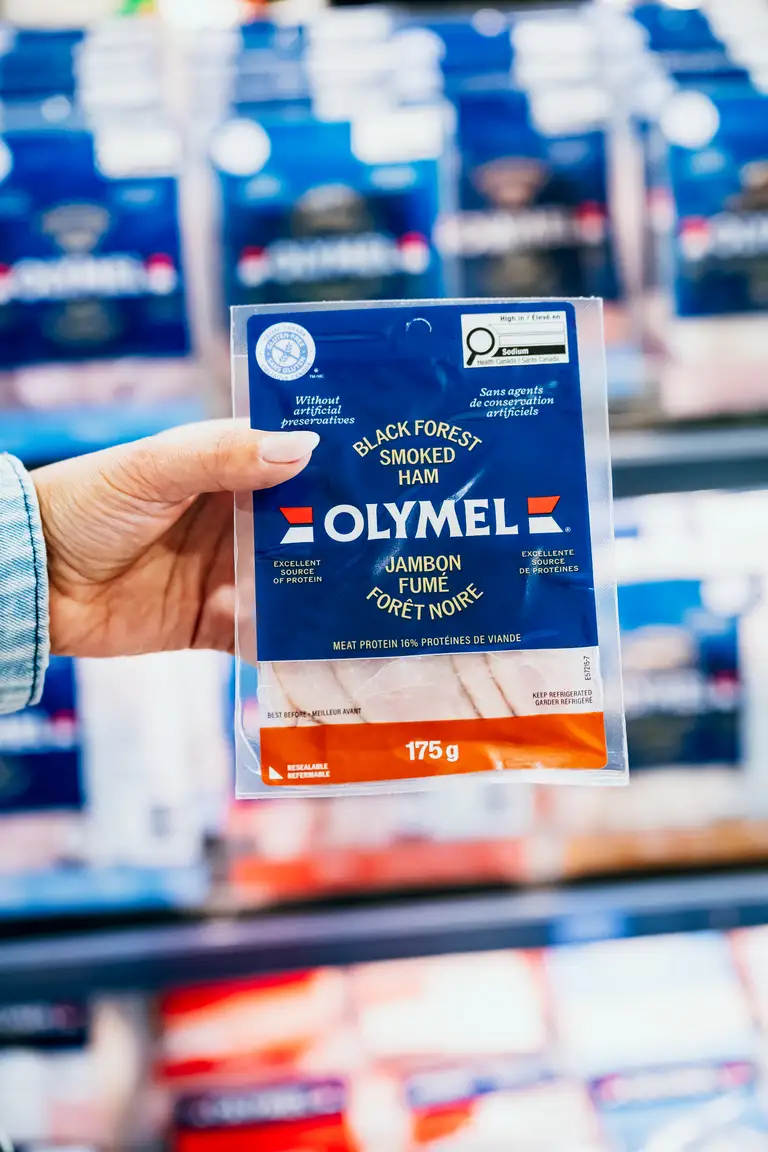 Une personne tenant un paquet de 175 g de jambon fumé Olymel Black Forest dans l'allée d'une épicerie, avec d'autres produits carnés en arrière-plan.