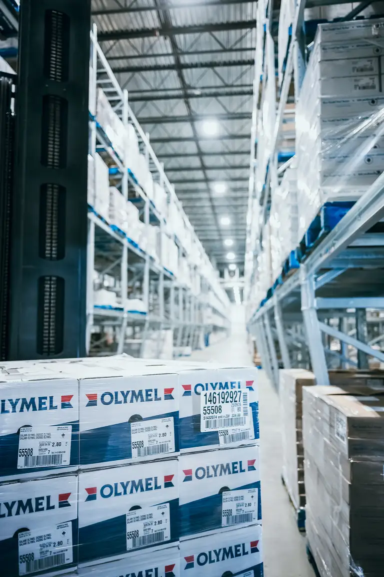 Intérieur d'entrepôt avec des boîtes empilées portant l'étiquette « Olymel » sur des palettes, des étagères métalliques et un haut plafond équipé de lampes fluorescentes.