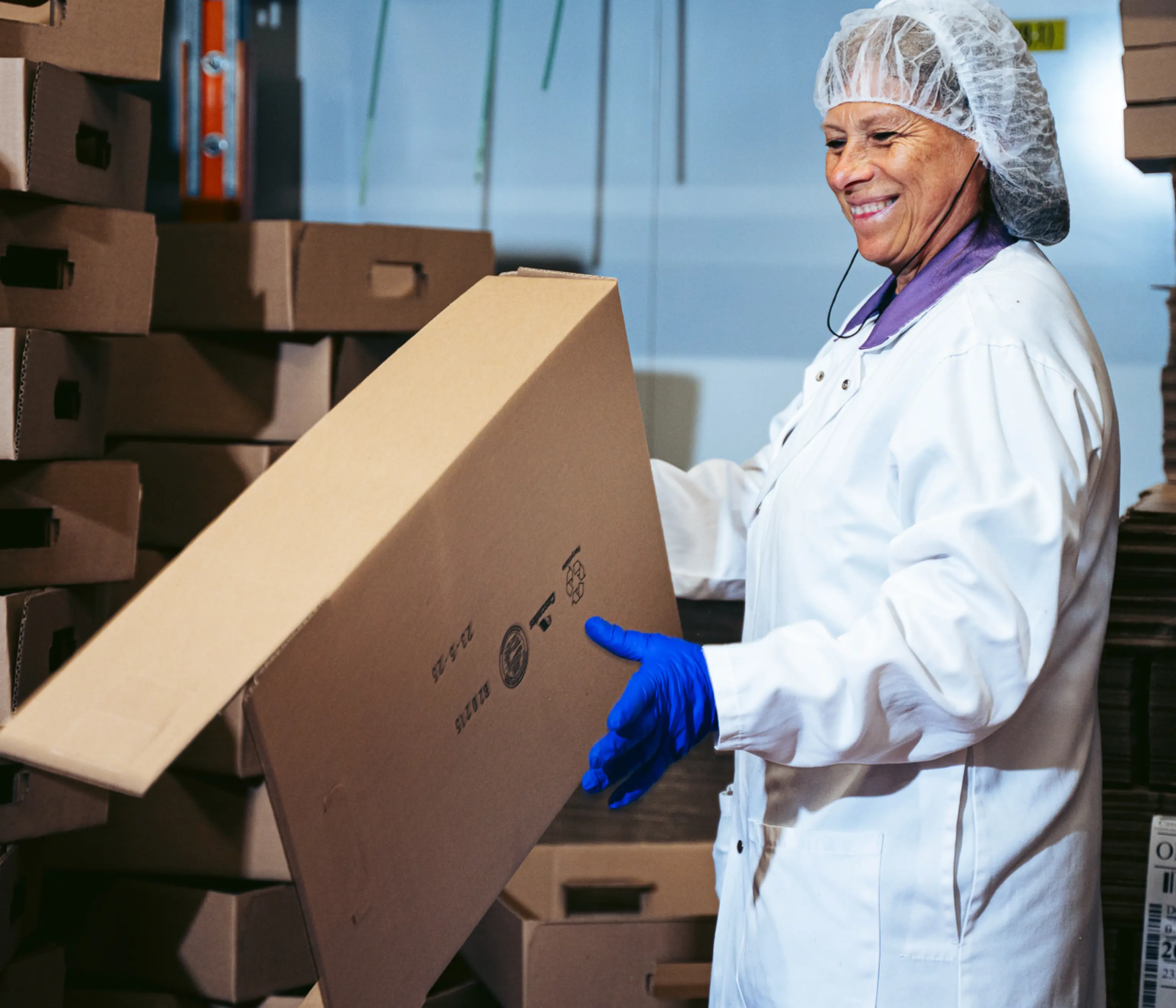 Un employé souriant, vêtu d'une blouse blanche et d'une charlotte, manipule des cartons dans un entrepôt, les mains gantées de bleu.