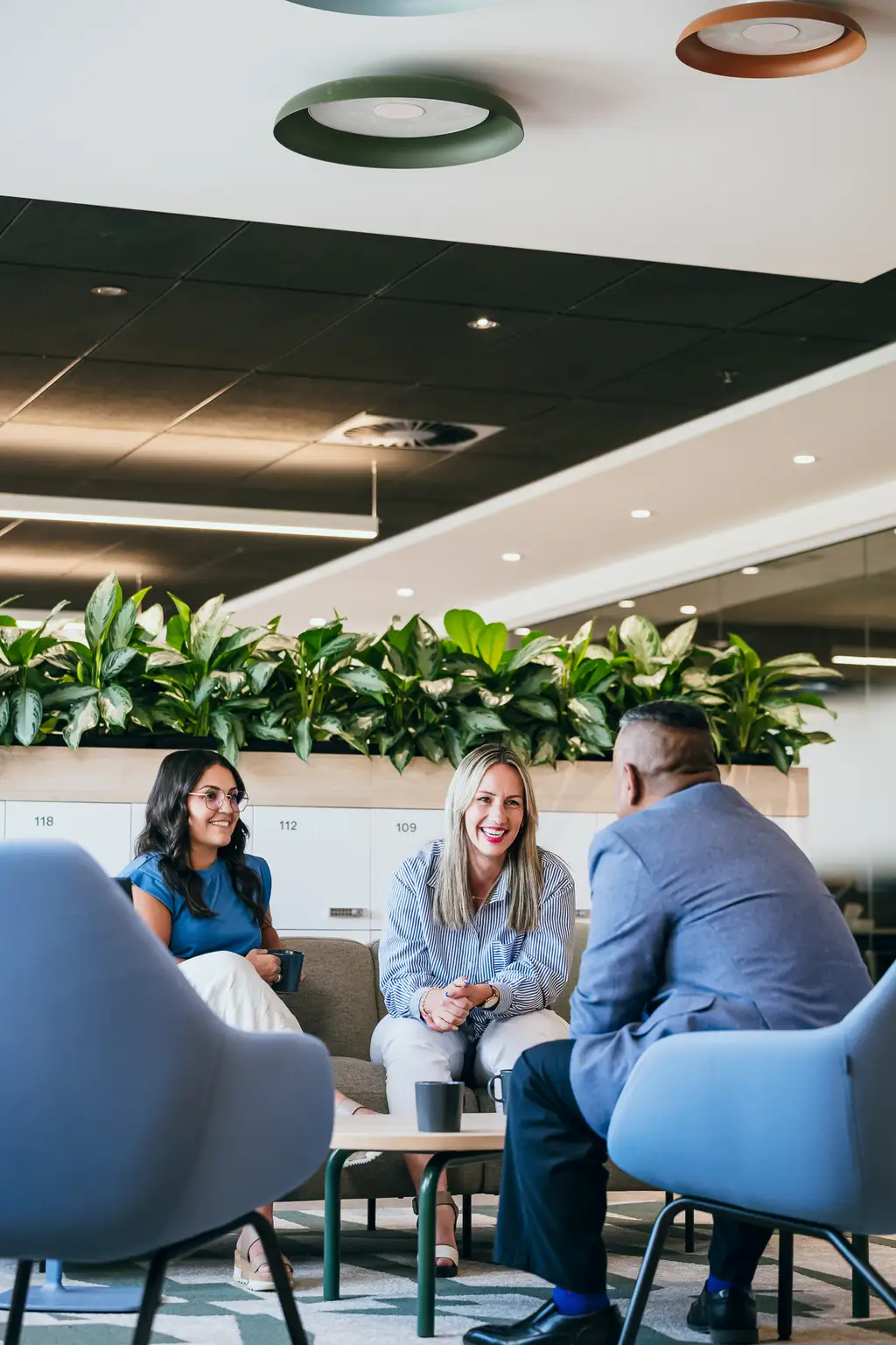 Trois personnes assises et discutant dans un salon de bureau moderne avec des plantes et des chaises élégantes.