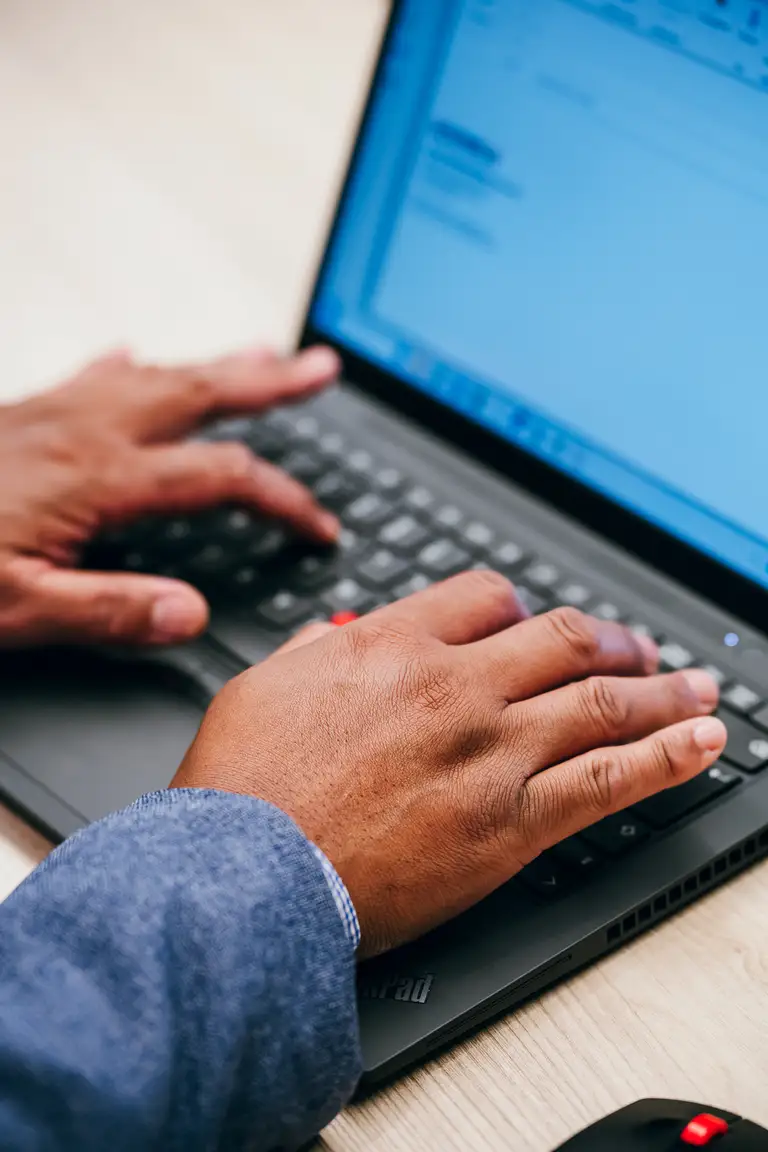 Personne tapant sur un ordinateur portable avec une interface de courriel visible, portant une veste bleue, assise à un bureau en bois clair.