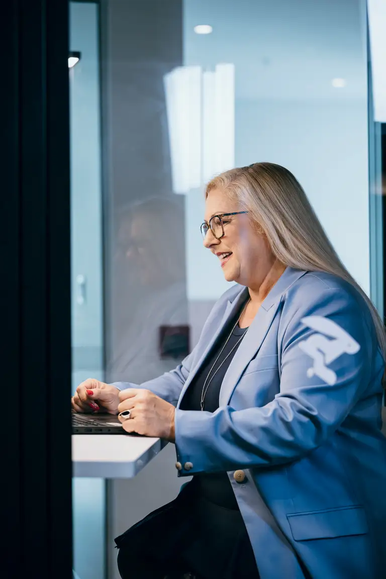 Une femme aux longs cheveux blonds et portant des lunettes, vêtue d'un blazer bleu, est assise à un bureau et utilise un ordinateur portable, souriant dans un environnement de bureau moderne.