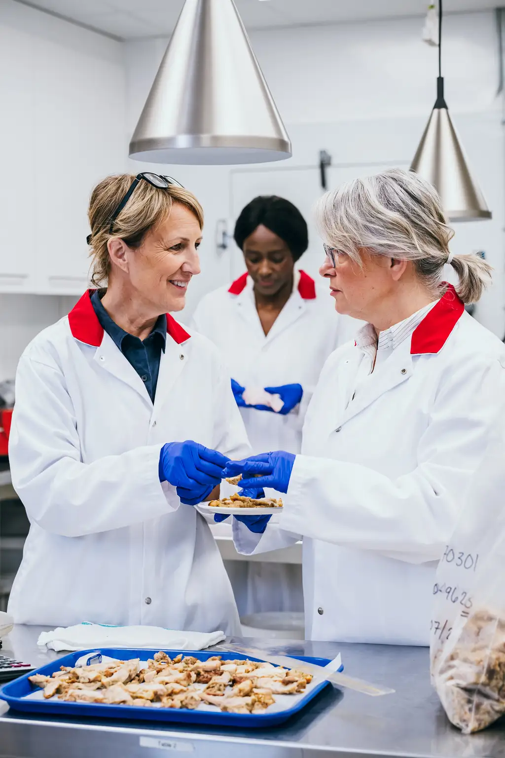 Trois scientifiques en blouse blanche et gants examinent des échantillons alimentaires sur des plateaux dans un laboratoire.