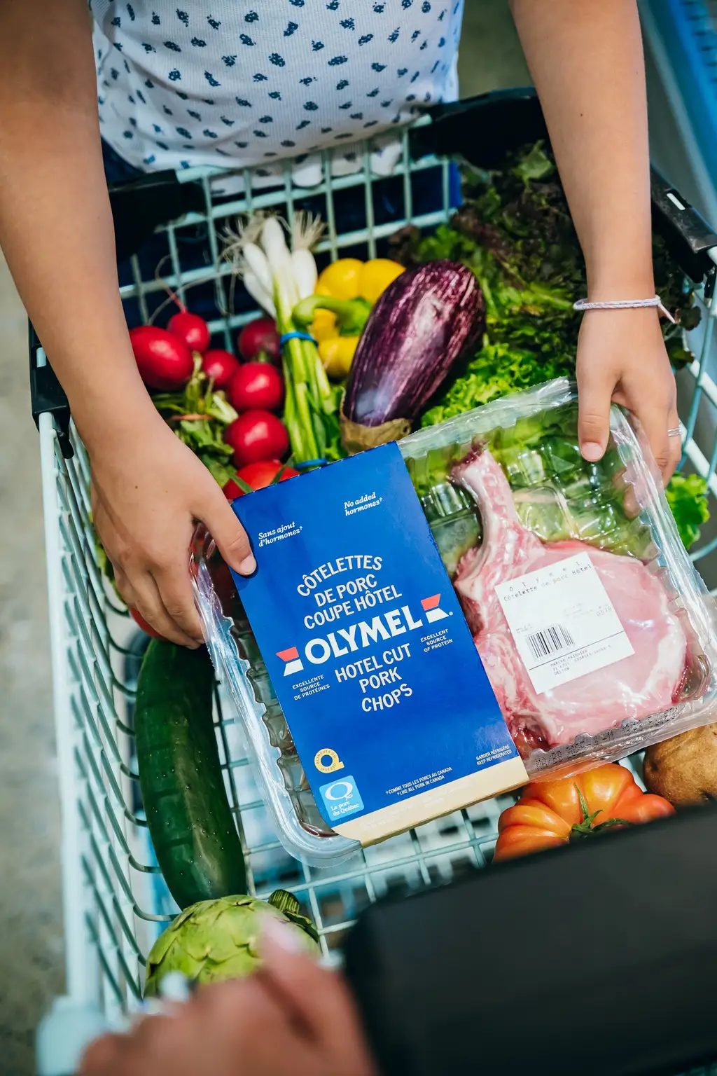 Enfant tenant un panier d’épicerie contenant des légumes frais, un paquet de côtelettes de porc et divers produits dans un magasin.