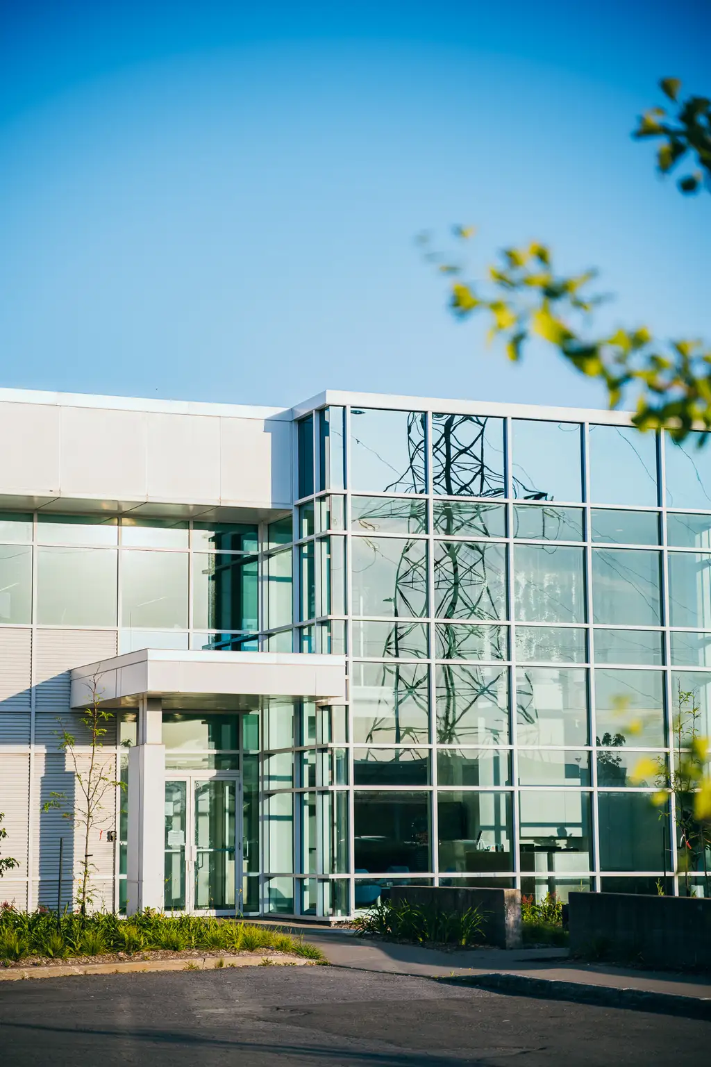 Bâtiment moderne en verre sous un ciel bleu clair, reflétant une tour électrique à proximité. De la verdure est visible au premier plan.