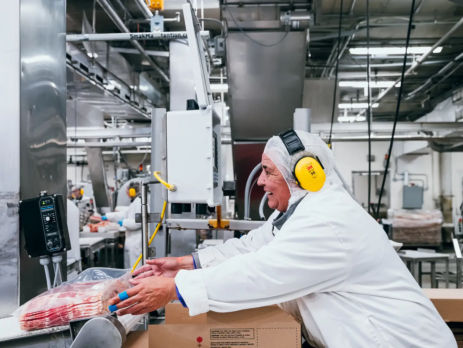Un ouvrier vêtu d'un équipement de protection et équipé d'une protection auditive emballe des articles dans une usine, avec des machines et des tapis roulants en arrière-plan.