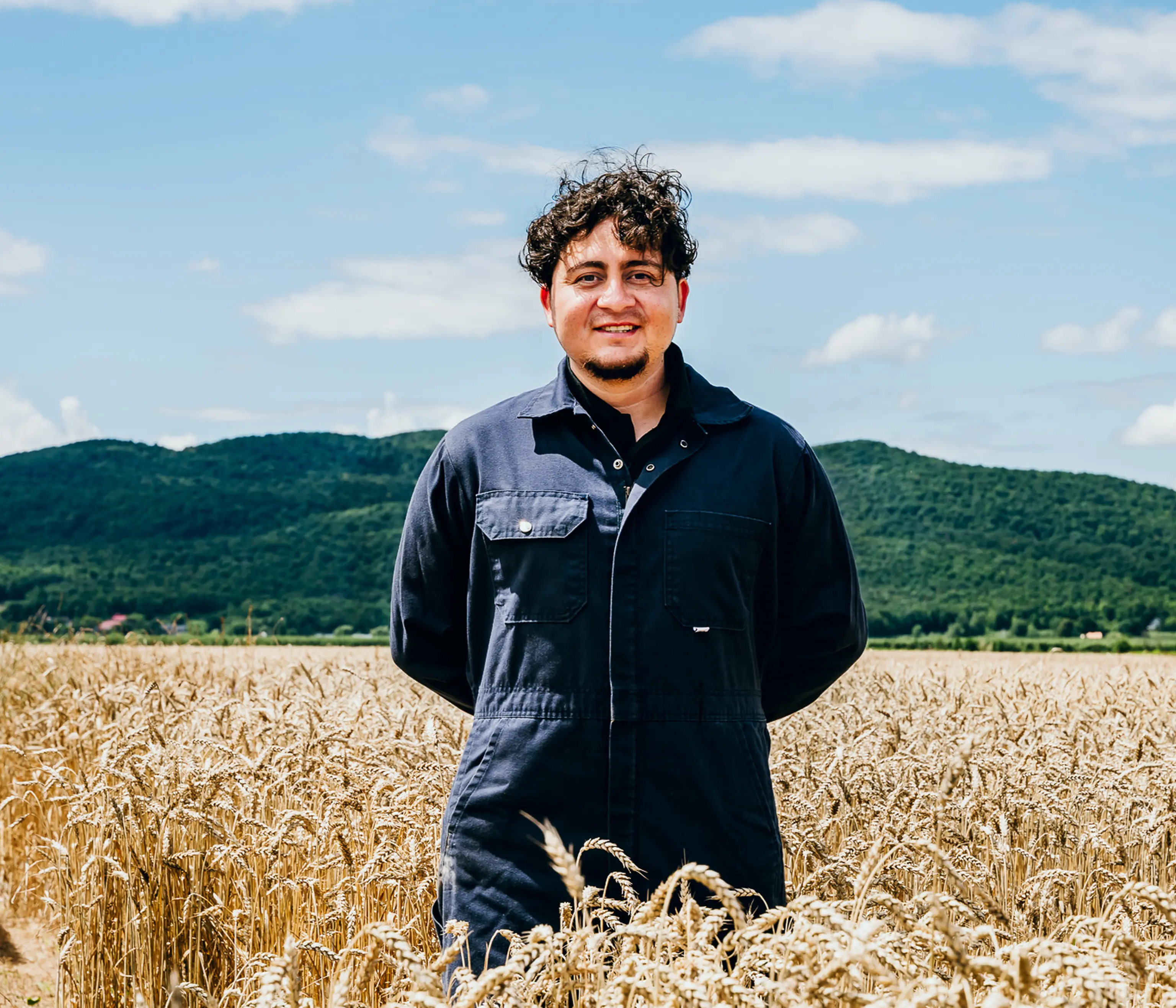 Une personne vêtue d'une combinaison bleue se tient debout, souriante, dans un champ de blé doré, avec des collines verdoyantes et un ciel bleu en arrière-plan.