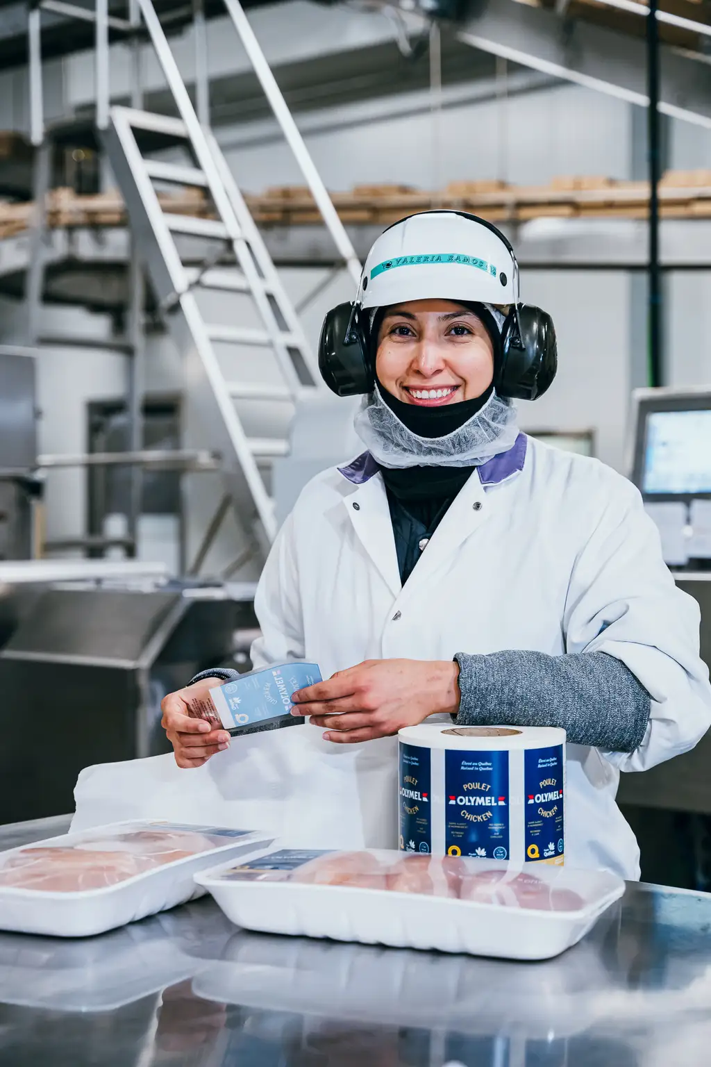 Un travailleur portant un équipement de protection sourit en manipulant de la viande emballée et des contenants d’assaisonnement dans une installation de transformation alimentaire.