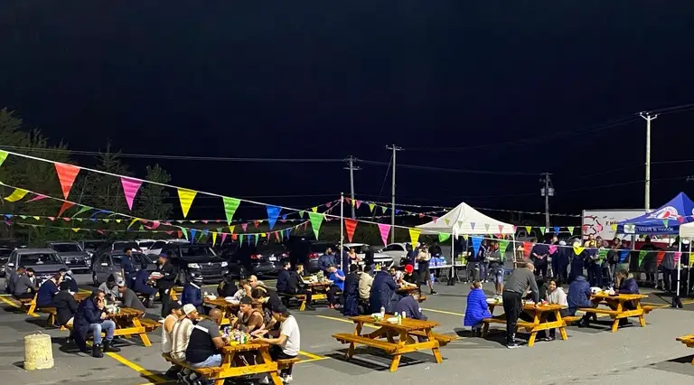 Des personnes assises à des tables de pique-nique sur un stationnement la nuit, entourées de fanions colorés et de tentes, avec des voitures stationnées en arrière-plan.