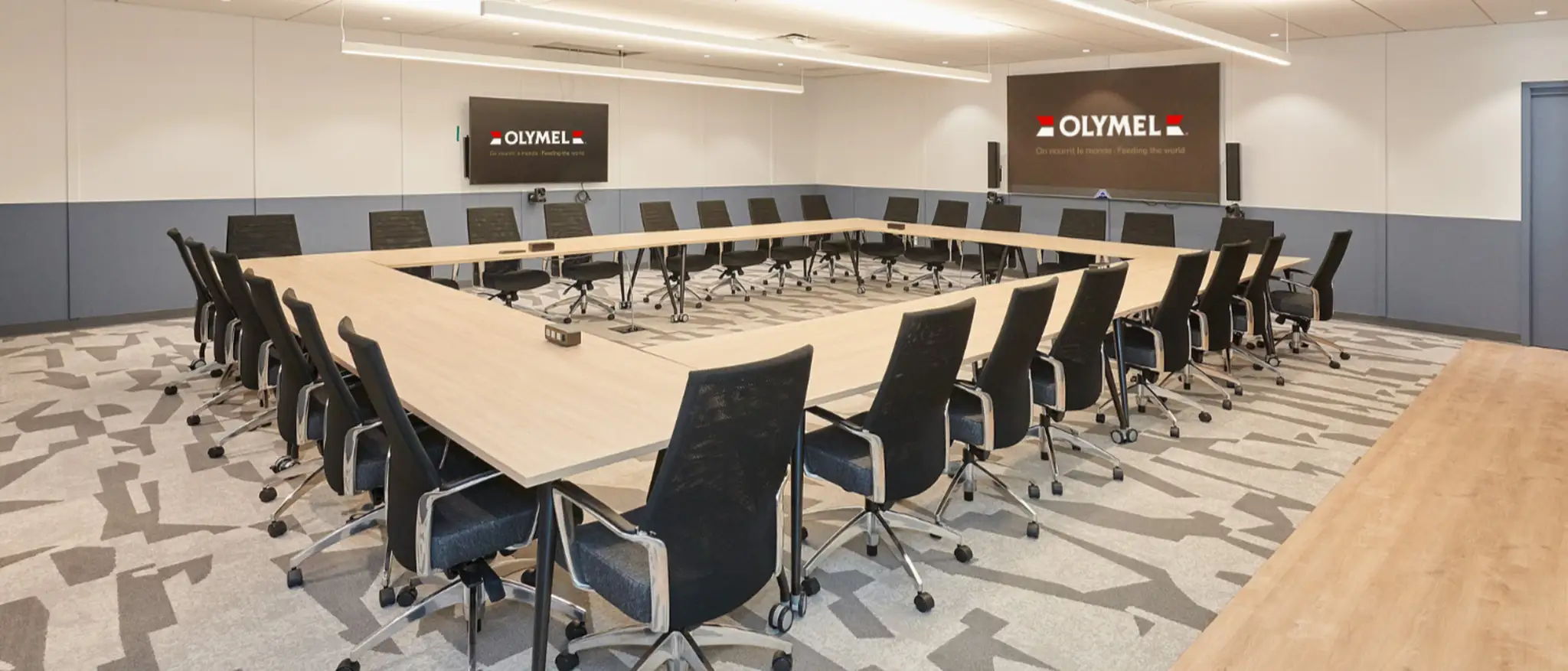 Une salle de conférence moderne avec une grande table en U, des chaises noires et des écrans affichant le logo d’Olymel sur les murs.