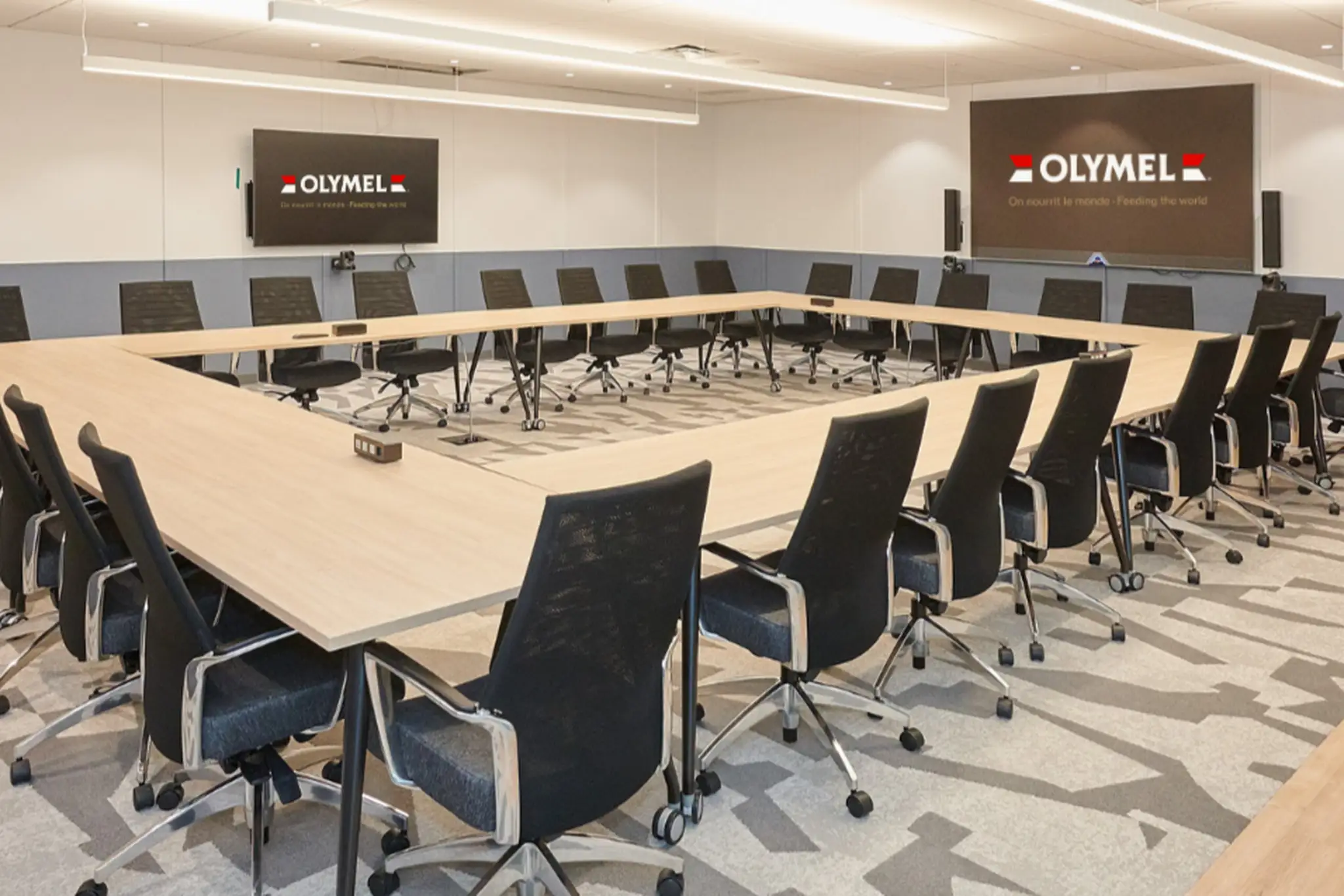 Une salle de conférence moderne avec une grande table en U, des chaises noires et des écrans affichant le logo d’Olymel sur les murs.
