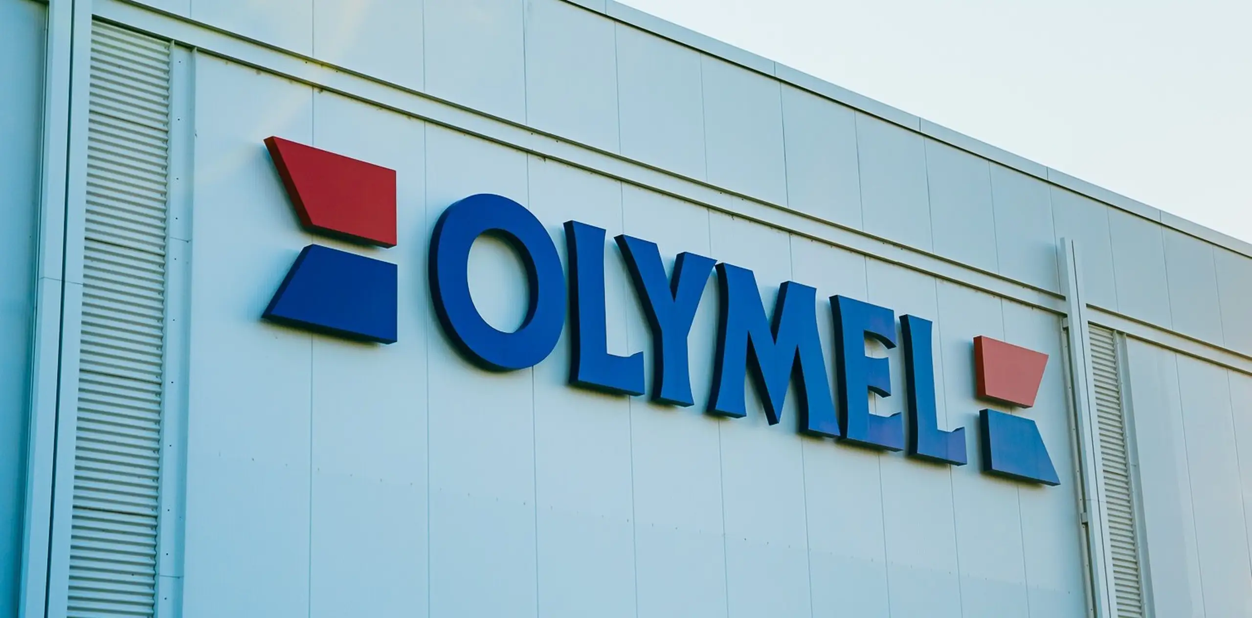Façade d’un bâtiment avec le logo d’Olymel en bleu et rouge, comportant des flèches stylisées, sur fond blanc.