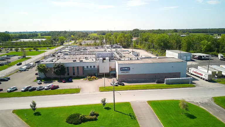 Vue aérienne d’un bâtiment industriel entouré de verdure, avec des voitures stationnées et des routes adjacentes sous un ciel bleu clair.