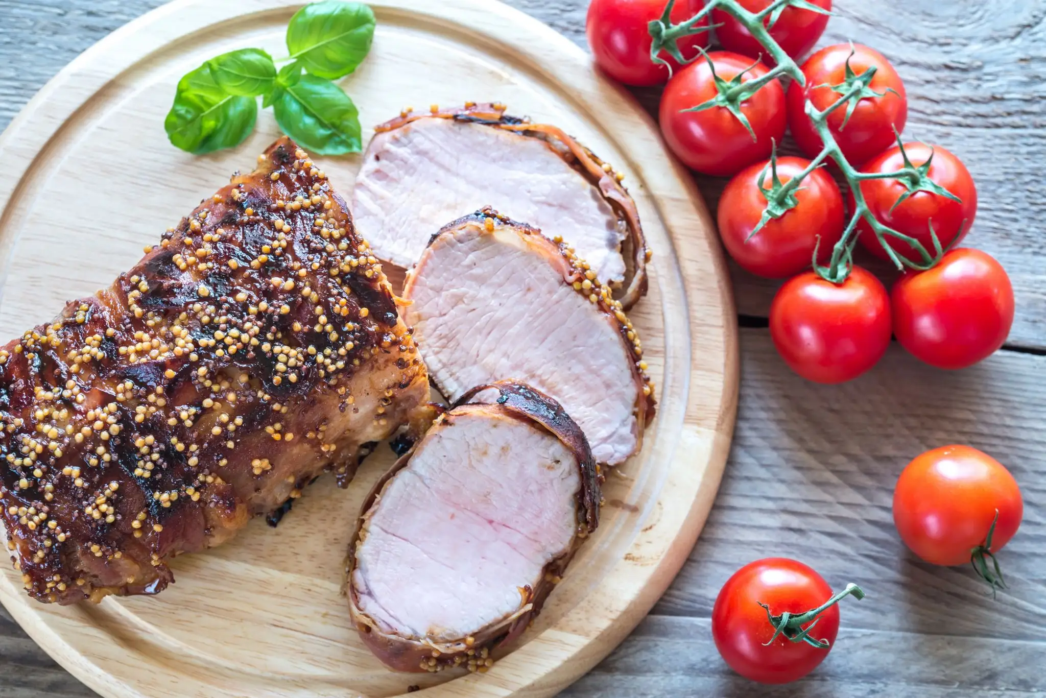 Rôti de porc tranché avec des graines de moutarde sur une planche en bois, garni de basilic, à côté d’une grappe de tomates fraîches sur vigne.
