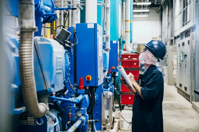 Un travailleur en tenue de protection examine une machine dans une installation industrielle, tenant une planchette à pince, entouré d’équipements et de tuyaux bleus.