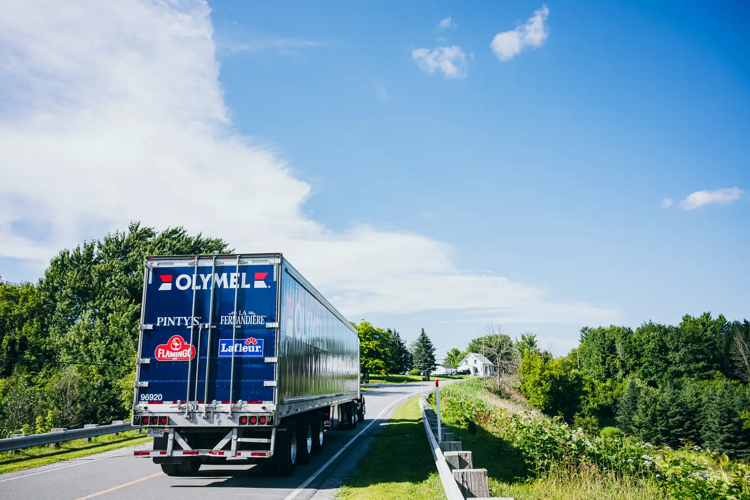 Un camion bleu d’Olymel circule sur une route rurale bordée de verdure, avec une maison blanche sous un ciel bleu clair.