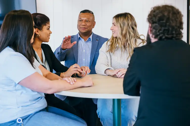 Un groupe de cinq personnes assises autour d'une table, engagées dans une discussion, dont une personne gesticule en parlant.