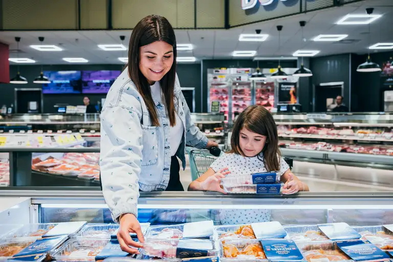 Une femme et une fille achètent des produits de la mer emballés dans une épicerie, en fouillant dans un congélateur. Des étagères de viande sont visibles à l'arrière-plan.