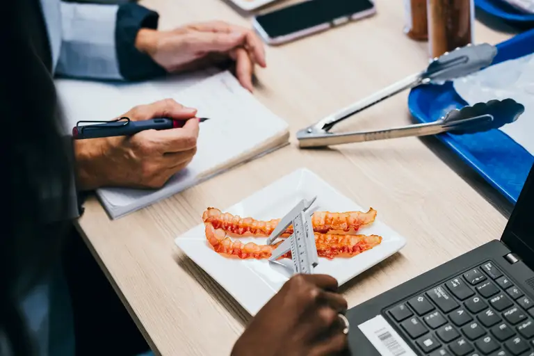 Personne mesurant des tranches de bacon avec un pied à coulisse sur une assiette, pendant qu’une autre prend des notes. À proximité, on trouve un ordinateur portable, une pince et un téléphone intelligent sur une table.