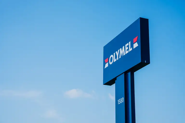Une enseigne inscrite «Olymel» se trouve devant un fond de ciel bleu parsemé de petits nuages.