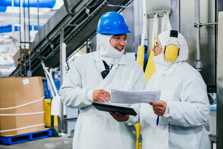 Deux ouvriers vêtus de combinaisons de protection et casqués discutent dans un environnement industriel, tenant une tablette et des documents.