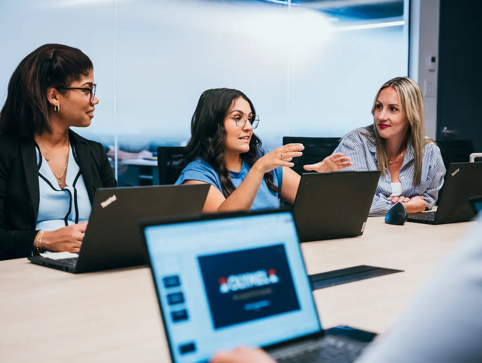Trois femmes discutent autour d'une table de conférence, équipées d'ordinateurs portables, dans un bureau moderne.