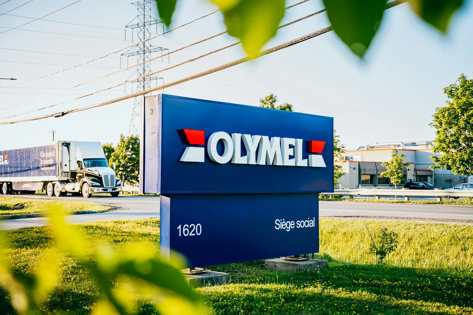 Enseigne indiquant "OLYMEL" avec un camion en arrière-plan, entourée de verdure et sous un ciel bleu clair.