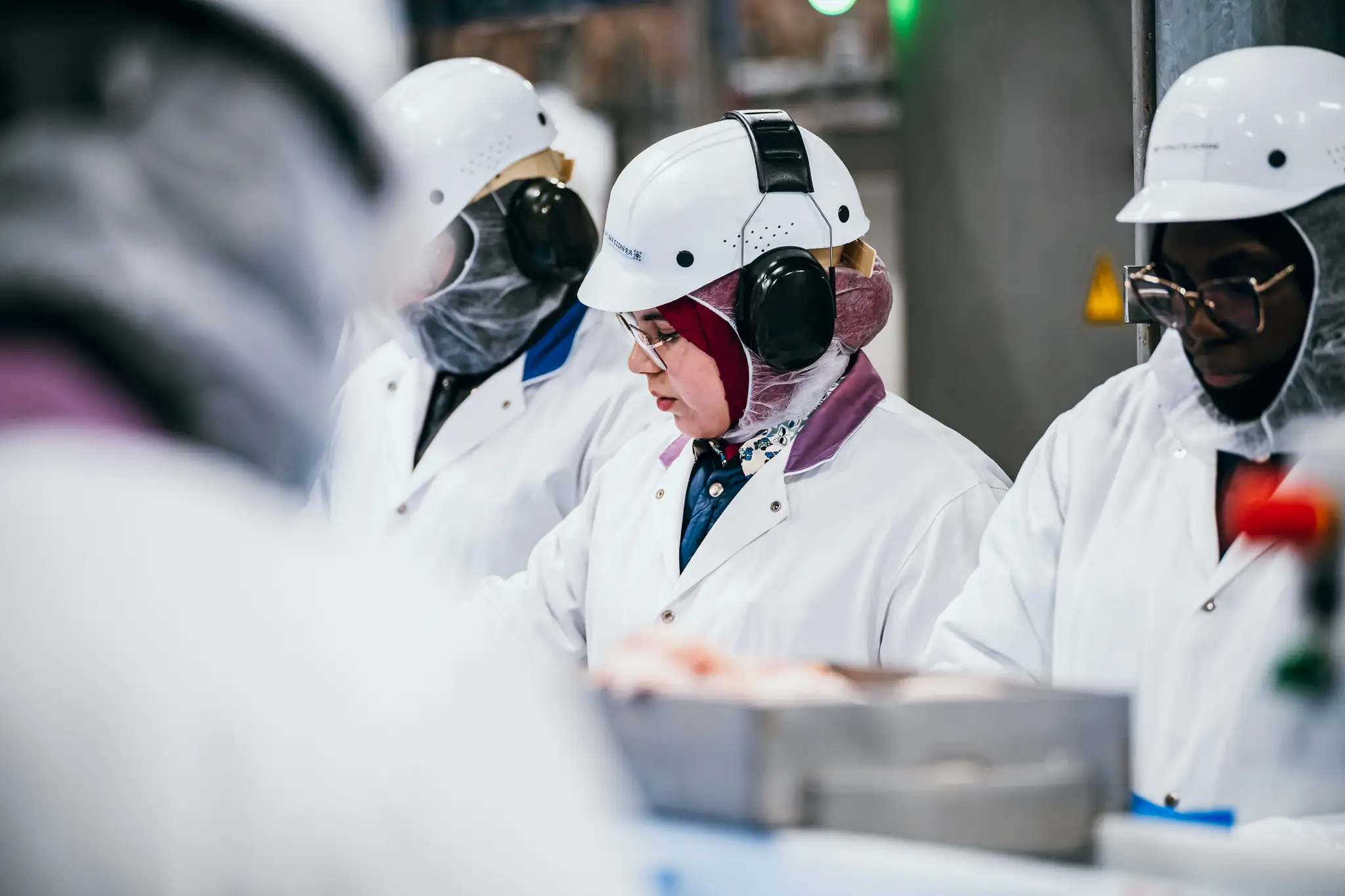 Des ouvriers vêtus de blouses blanches, casqués et équipés de protections auditives se concentrent sur leurs tâches à la chaîne de production dans un environnement industriel.