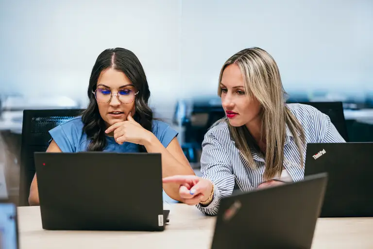 Deux femmes travaillent ensemble sur des ordinateurs portables dans un bureau, l'une pointant vers un écran pendant que l'autre regarde pensivement.