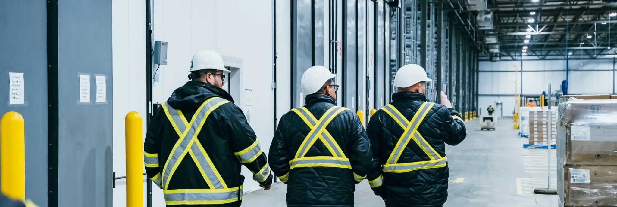 Trois travailleurs portant des équipements de sécurité et des casques marchent dans une grande installation industrielle, avec de hauts plafonds et des palettes empilées.