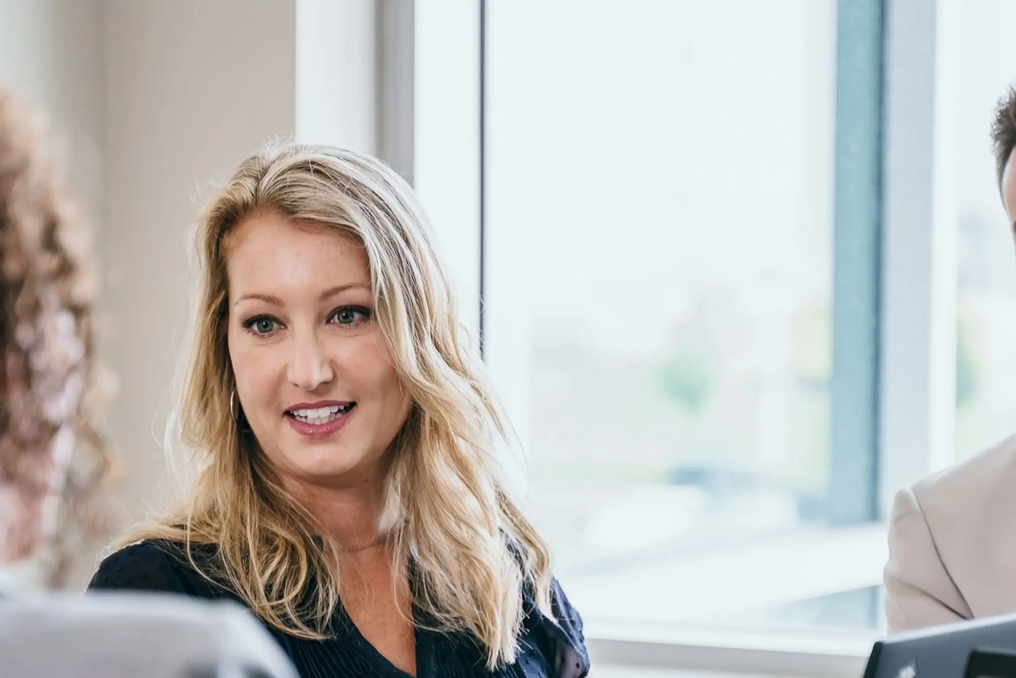Trois personnes en réunion d’affaires, assises autour d’une table avec des ordinateurs portables, en pleine conversation près d’une fenêtre.
