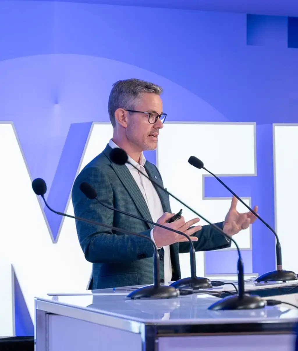Yanick Gervais en complet, s’exprimant à un podium avec des micros, faisant des gestes avec les mains, devant une grande enseigne illuminée sur fond bleu.
